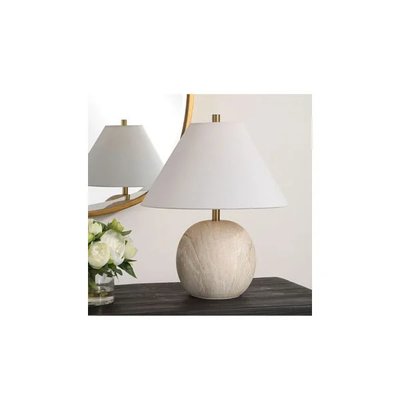 Lampe de table élégante avec base sphérique en pierre texturée beige et abat-jour conique blanc, posée sur une table en bois sombre à côté d’un vase de fleurs blanches et d’un miroir rond doré, image provenant de Royaume Luminaire