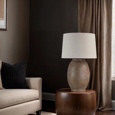 Lampe de table au pied en céramique finition pierre brune et abat-jour cylindrique beige, posée sur une table d’appoint ronde en bois, à côté d’un canapé crème avec coussins décoratifs, dans un salon aux murs foncés et rideaux texturés, image provenant de Linen Chest