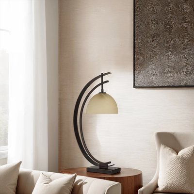 Lampe de table au design moderne avec base rectangulaire noire et structure arquée en métal soutenant un abat-jour hémisphérique beige, posée sur une table d’appoint ronde en bois, à côté d’un canapé crème et d’un fauteuil beige, devant un mur clair décoré d’un tableau texturé, image provenant de Linen Chest