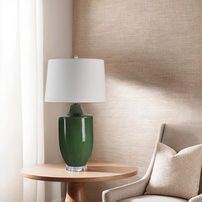 Lampe de table avec base en céramique émaillée vert foncé et abat-jour cylindrique blanc, posée sur une table ronde en bois clair à côté d’un fauteuil beige avec coussin texturé, devant un mur beige texturé, image provenant de Linen Chest