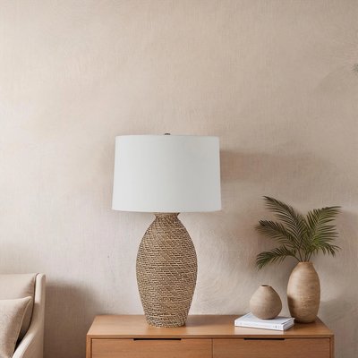 Lampe de table avec base en céramique imitation rotin tressé et abat-jour cylindrique blanc, posée sur un meuble en bois clair à côté de vases décoratifs et d’une petite plante verte, devant un mur texturé beige, image provenant de Linen Chest