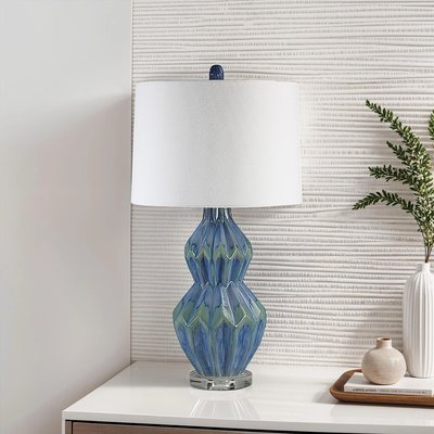 Lampe de table élégante avec base en verre bleu facetté aux reflets verts et design géométrique, surmontée d’un abat-jour cylindrique blanc, posée sur un meuble blanc avec plateau décoratif et vases, devant un mur texturé clair, image provenant de Linen Chest