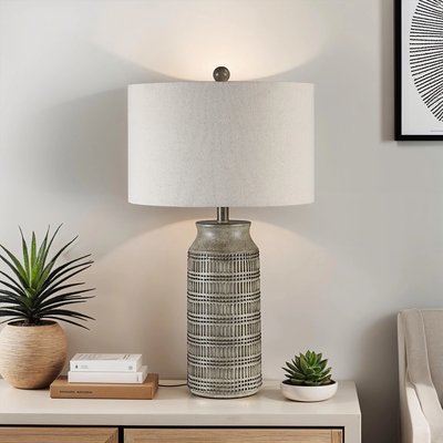 Lampe de table avec base en céramique grise ornée de motifs géométriques en relief et abat-jour cylindrique en tissu beige, posée sur une table en bois clair avec deux petites plantes décoratives et des livres, image provenant de Linen Chest