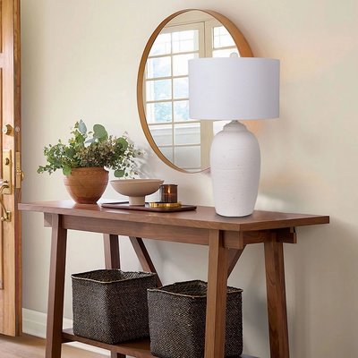 Lampe de table au pied en céramique blanche texturée et abat-jour cylindrique blanc, posée sur une console en bois dans une entrée décorée d’un miroir rond, de plantes en pot et de paniers de rangement, image provenant de Linen Chest