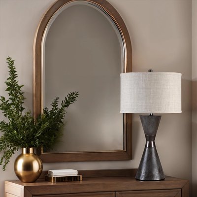 Lampe de table au design moderne avec base en forme de sablier en métal brun foncé et abat-jour cylindrique en tissu beige clair, posée sur une commode en bois devant un grand miroir à cadre arqué, accompagnée d’un vase doré contenant un feuillage vert, image provenant de Linen Chest