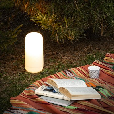 Lampe de table cylindrique blanche utilisée en extérieur sur une couverture colorée avec livres et tasse, photo par Luminaires & cie