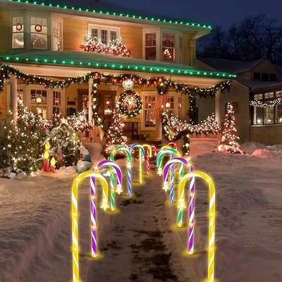 Maison décorée pour Noël avec des guirlandes lumineuses, un sapin, et une allée bordée de cannes de bonbon lumineuses multicolores plantées dans la neige - Amazon