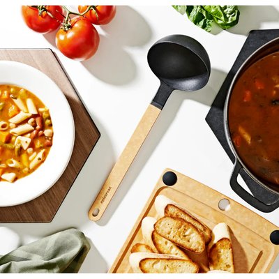 Louche noire au manche en bois naturel de la marque Epicurean, posée sur une table blanche entre une soupe minestrone, une planche de présentation avec crostinis et une marmite de soupe – accessoire de cuisine élégant et fonctionnel offert chez La Guilde Culinaire