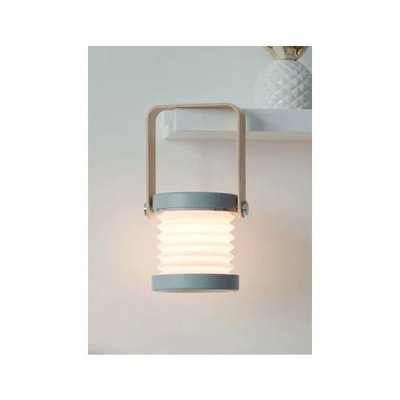 Lampe de table portable en forme de lanterne avec structure en bois clair, bordures grises et diffuseur plissé blanc diffusant une lumière douce, posée sur une étagère, image provenant de Royaume Luminaire