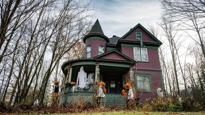 Grande maison victorienne décorée pour l'Halloween avec des fantômes suspendus, des squelettes, des citrouilles illuminées et des décorations automnales dans un cadre boisé.