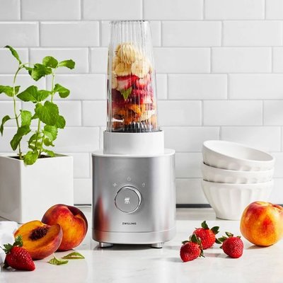 Mélangeur individuel ZWILLING Enfinigy en inox brossé avec contenant transparent rempli de fruits frais – fraises, bananes et pêches – sur un comptoir de cuisine moderne, chez Linen Chest