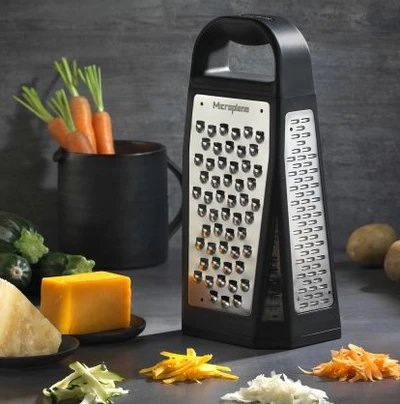 Râpe 4 côtés Elite Microplane posée sur un comptoir de cuisine avec légumes râpés, fromage et carottes – Accessoire de cuisine polyvalent disponible à La Guilde Culinaire