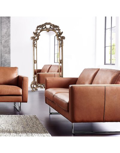 Grand miroir sur pied à cadre ornementé doré de chez Ô Miroir, placé dans un salon contemporain avec canapés en cuir brun.
