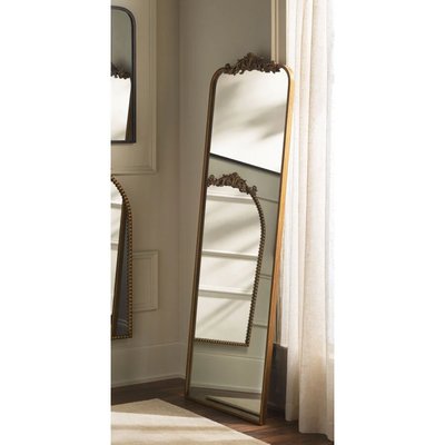 Grand miroir sur pied style antique avec cadre doré orné, provenant de Lovaco, placé près d’une fenêtre avec rideaux clairs.