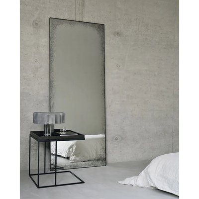 Miroir rectangulaire sur pied avec cadre noir et effet vieilli de Lovaco, placé contre un mur en béton dans une chambre minimaliste.