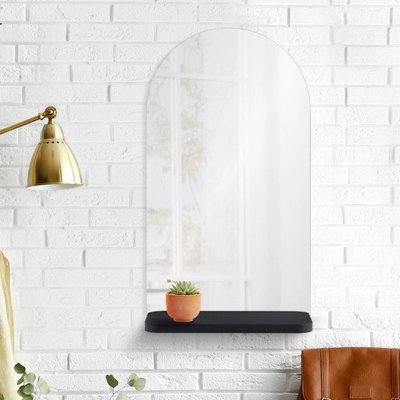 Miroir Lucila chez Linen Chest – Miroir mural en forme d’arche avec tablette noire intégrée, fixé sur un mur en briques blanches et décoré d’une petite plante en pot.