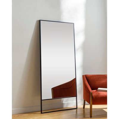 Miroir pleine longueur à cadre noir minimaliste de Lovaco, appuyé contre un mur blanc près d’un fauteuil orange moderne.