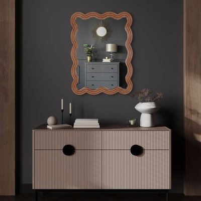 Miroir mural Adria vertical chez Linen Chest – Miroir décoratif en bois aux contours ondulés, accroché sur un mur gris foncé au-dessus d’une commode moderne aux lignes épurées.