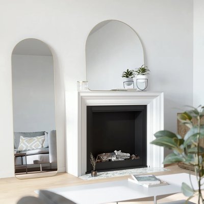 Deux miroirs arrondis à cadre doré de chez Kozy, l’un mural et l’autre sur pied, décorant un salon lumineux avec cheminée blanche et plantes vertes.