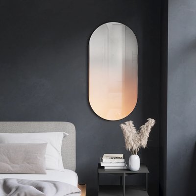 Miroir mural ovale avec effet dégradé cuivre de chez Kozy, accroché sur un mur foncé dans une chambre moderne avec table de chevet minimaliste.