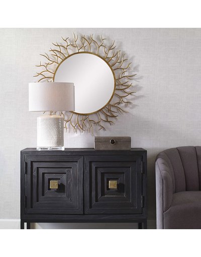 Miroir rond style soleil à cadre doré de chez Ô Miroir, accroché au-dessus d’un buffet noir sculpté avec lampe blanche et fauteuil gris.