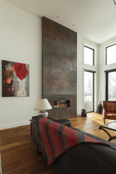 Salon contemporain avec foyer mural encastré dans un revêtement effet béton vieilli, grandes fenêtres laissant entrer une lumière naturelle abondante, plancher de bois franc chaleureux, fauteuil à motifs écossais, canapé en cuir foncé avec jeté rouge, lampe décorative sur table d’appoint et tableau artistique abstrait accroché au mur blanc de chez Mur Tendance.