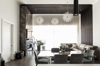 Salle de séjour moderne et élégante avec plafond en lattes de bois foncé, luminaires suspendus en forme de sphères florales, mur accent noir texturé, canapé sectionnel en cuir brun, table à manger en verre avec chaises grises en velours, mur de marbre sombre avec foyer encastré et télévision, grandes fenêtres avec voilages blancs laissant entrer la lumière naturelle de chez Mur Tendance.
