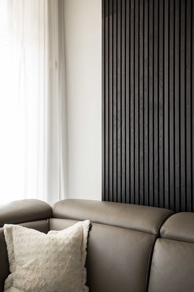 Mur accent en lattes de bois noir verticales dans un salon moderne, juxtaposé à un mur blanc avec rideau translucide, canapé en cuir taupe et coussin crème texturé, pour une ambiance chic et épurée de chez Mur Tendance.