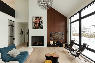 Salon contemporain lumineux avec plafond cathédrale, mur accent en lattes de bois brun foncé, grandes fenêtres pleine hauteur avec vue sur l’extérieur enneigé, foyer moderne encastré, œuvres d’art murales, mobilier design incluant canapé bicolore, fauteuils en cuir noir et tables basses rondes en bois naturel et velours jaune moutarde de chez Mur Tendance.