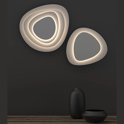 Murale décorative lumineuse Abstract Panels de chez Luminaires & Cie composée de deux formes organiques superposées, émettant une lumière douce et diffuse sur un mur foncé, avec un vase noir mat et un bol assorti placés sur une surface en bois