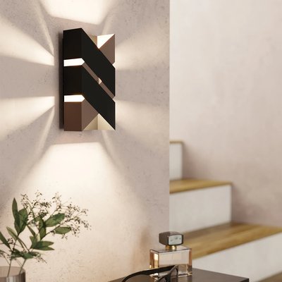 Applique murale moderne Gurare de chez Luminaires & Cie avec design géométrique noir et bronze, diffusant une lumière indirecte vers le haut et le bas, installée dans un intérieur contemporain aux murs pâles près d'un escalier en bois et d'une table décorative
