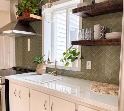 Cuisine lumineuse de style rustique-chic avec dosseret en céramique verte posée en chevrons, comptoir en quartz blanc lustré, robinet doré vintage, étagères en bois massif flottantes garnies de vaisselle, plantes décoratives et évier encastré sous une grande fenêtre apportant une belle lumière naturelle de chez Muraluxe