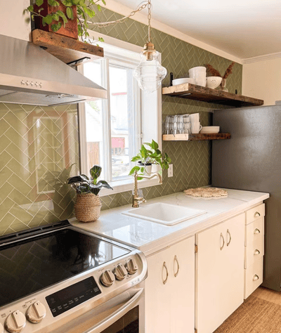Cuisine chaleureuse avec dosseret en tuiles vert olive en motif chevron, comptoir blanc lustré, armoires inférieures crème avec poignées dorées, cuisinière moderne en inox, robinet doré, étagères murales en bois foncé décorées de vaisselle blanche et de plantes, le tout éclairé par une grande fenêtre apportant une belle lumière naturelle de chez Muraluxe