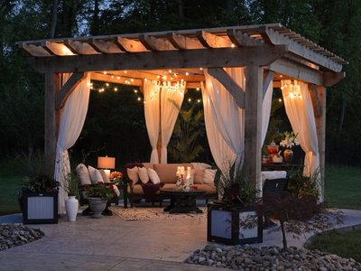 Pergola par Dengarden – Pergola en bois avec rideaux blancs et guirlandes lumineuses, aménagée avec un salon extérieur chaleureux et des plantes décoratives, parfaite pour une soirée conviviale dans le jardin.