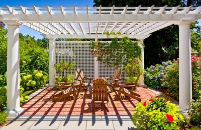 Pergola blanche avec colonnes classiques et treillis décoratif ombrageant un coin détente avec chaises en bois, entouré de fleurs colorées, provenant de Decoist.