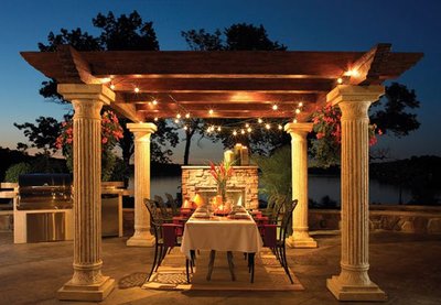 Pergola extérieure avec colonnes classiques éclairée de guirlandes lumineuses et table dressée pour un souper au coucher du soleil, provenant de Decoist.