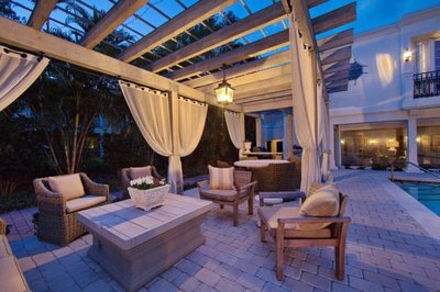 Pergola élégante en bois clair avec rideaux blancs et mobilier de jardin confortable près d’une piscine, provenant de Decoist.