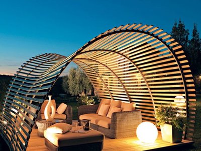 Pergola moderne en bois aux courbes architecturales abritant un coin lounge extérieur avec mobilier en rotin et luminaires sphériques, provenant de Decoist.