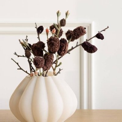 Arrangement floral minimaliste avec branches et fleurs séchées brun foncé dans un vase blanc mat ondulé, posé sur une table en bois clair devant un mur neutre chez décors véronneau