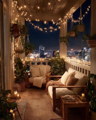 Balcon de style bohème illuminé de guirlandes lumineuses Source - sandra novak Pinterest Balcon de style bohème illuminé de guirlandes lumineuses, meubles en bois avec coussins beiges, plantes suspendues et vue nocturne sur la ville scintillante Source - sandra novak Pinterest