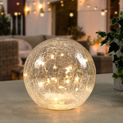 Lampe décorative en forme de sphère en verre craquelé, illuminée par de petites lumières LED chaudes, posée sur une table de terrasse avec un décor extérieur chaleureux en arrière-plan, image provenant de Linen Chest