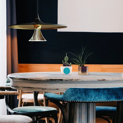 Suspension Ipno au design futuriste en métal doré suspendue au-dessus d'une table ronde vintage en bois patiné, entourée de chaises rembourrées en velours bleu, dans une salle à manger éclectique et chaleureuse