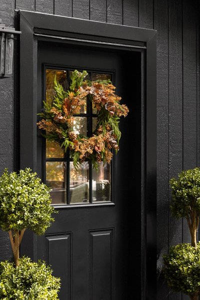 Couronne d'automne accrochée à une porte noire avec fenêtres, composée de feuillage vert, branches orangées et baies décoratives, encadrée par deux arbustes taillés symétriquement par Décors Véronneau
