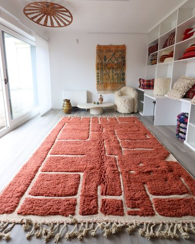 Pièce lumineuse avec grand tapis berbère orange à motifs géométriques, étagères de coussins colorés, fauteuil en laine bouclée et décoration murale artisanale Source - Tree of Colors Pinterest