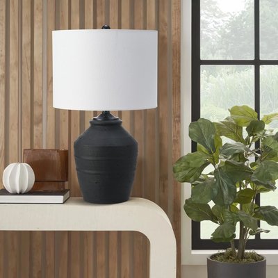 Lampe de table avec pied en céramique noir mat au design artisanal et abat-jour cylindrique blanc, posée sur une console blanche incurvée, accompagnée de décorations modernes et d’une plante verte en pot près d’une fenêtre, image provenant de Linen Chest