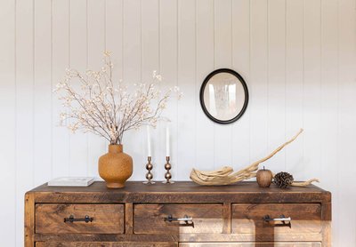 Commode en bois rustique décorée avec un vase terracotta et branches fleuries, chandeliers en métal, éléments naturels et petit miroir rond sur mur à panneaux blancs par Décors Véronneau