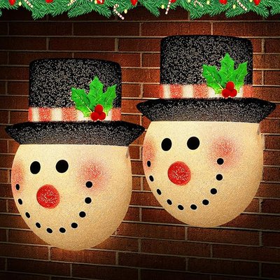 Deux décorations lumineuses murales en forme de visages de bonhommes de neige avec chapeaux noirs à paillettes, ornés de houx, accrochées sur un mur de briques avec une guirlande de Noël en haut par AMAZON