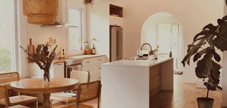 Cuisine lumineuse avec grand îlot en quartz signé Caroline Beaulieu spécialiste en design d'intérieur Cuisine lumineuse avec grand îlot en quartz, armoires blanches, accents en bois et coin repas avec table ronde en bois et chaises en rotin signé Caroline Beaulieu spécialiste en design d'intérieur