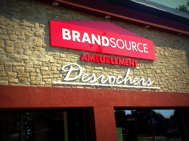 Enseigne extérieure de la boutique Ameublement BrandSource Desrochers Enseigne extérieure de la boutique Ameublement BrandSource Desrochers sur un mur de pierre, avec le logo rouge BrandSource