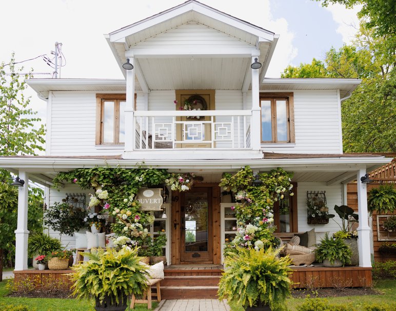 Façade extérieure fleurie de La Maison de Margo, une charmante boutique de décoration située dans une maison blanche aux volets en bois, avec véranda accueillante, végétation abondante et ambiance chaleureuse
